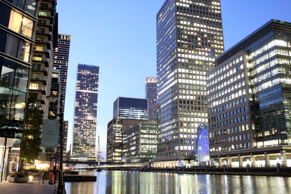 Photograph of modern high-rise office buildings in Canary Wharf, with glass facades reflecting the evening sky. The buildings are illuminated with interior and exterior lighting, showcasing a combination of reflective glass and steel surfaces. The scene features a canal running alongside the buildings, with calm water reflecting the structures above. The lighting creates a bright and vibrant atmosphere, typical of a bustling commercial district. Canary Wharf Carpet Cleaning is not visible but is mentioned as the service provider for cleaning within this context of deep cleaning and surface maintenance in a professional environment.