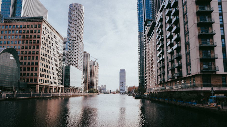 A wide view of Canary Wharf in London featuring tall modern skyscrapers with glass and steel facades lining a calm river. The buildings vary in architectural styles, including curved and rectangular shapes, with some under construction visible in the background. The sky is partly cloudy, and the water reflects the cityscape, contributing to a clean, urban atmosphere. The scene is well-lit, emphasizing the clarity and sharpness of the commercial district, which is associated with professional cleaning and surface maintenance services provided by Canary Wharf Carpet Cleaning.