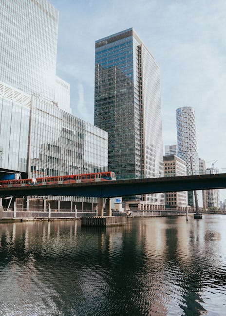 Photograph of modern high-rise office buildings in Canary Wharf, with glass facades reflecting the evening sky. The buildings are illuminated with interior and exterior lighting, showcasing a combination of reflective glass and steel surfaces. The scene features a canal running alongside the buildings, with calm water reflecting the structures above. The lighting creates a bright and vibrant atmosphere, typical of a bustling commercial district. Canary Wharf Carpet Cleaning is not visible but is mentioned as the service provider for cleaning within this context of deep cleaning and surface maintenance in a professional environment.