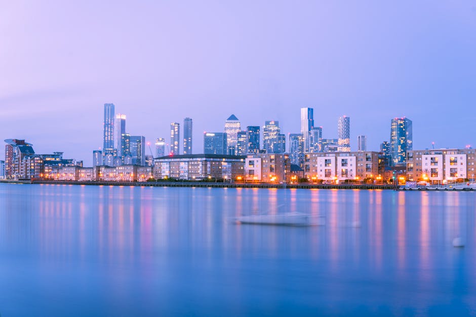 A panoramic view of the Canary Wharf skyline taken during twilight, showcasing modern glass skyscrapers, office buildings, and residential apartments reflecting on the calm river below. The scene features well-lit high-rise towers with illuminated windows and exterior lighting, set against a soft purple and blue evening sky. The foreground includes the tranquil water with gentle ripples, and the cityscape is bordered by low-rise residential buildings along the waterfront. This image exemplifies urban surface cleaning and maintenance in a vibrant commercial district, as captured by Canary Wharf Carpet Cleaning.