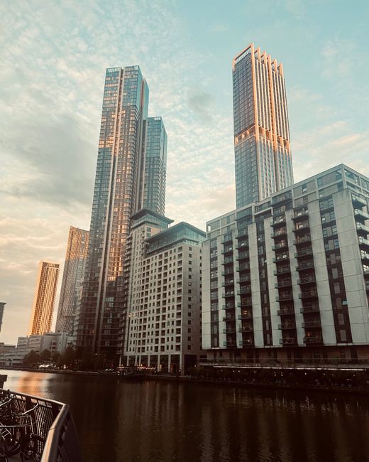 A high-rise urban scene in Canary Wharf with several modern glass and steel skyscrapers reflecting the sky during sunset. The tallest building features illuminated vertical accents, and the lower floors consist of commercial and residential units with numerous balconies. In the foreground, a calm body of water mirrors the buildings, and part of a railing is visible at the bottom left corner. The lighting creates a warm glow on the building facades, emphasizing their sleek surfaces and architectural details. This image is associated with Heron Quays end of tenancy cleaning for Canary Wharf flats, provided by Canary Wharf Carpet Cleaning, showcasing a clean, professional environment suited for surface and deep cleaning services.