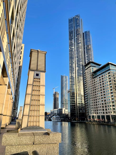 A panoramic view of the Canary Wharf skyline taken during twilight, showcasing modern glass skyscrapers, office buildings, and residential apartments reflecting on the calm river below. The scene features well-lit high-rise towers with illuminated windows and exterior lighting, set against a soft purple and blue evening sky. The foreground includes the tranquil water with gentle ripples, and the cityscape is bordered by low-rise residential buildings along the waterfront. This image exemplifies urban surface cleaning and maintenance in a vibrant commercial district, as captured by Canary Wharf Carpet Cleaning.