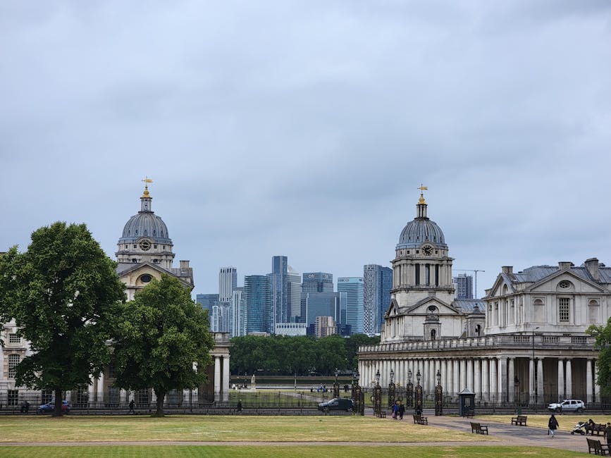 A wide view of Jubilee Park near Canary Wharf station showing historic neoclassical buildings with domed rooftops and ornate details, surrounded by green trees and open grassy areas. In the background, a dense cluster of modern skyscrapers with glass facades and rectangular shapes rise under a cloudy sky, creating a contrast between historic and contemporary architecture. The scene is outdoors during daylight, with some people walking or sitting on benches across the park, and a few vehicles visible near the buildings. The environment appears clean and well-maintained, typical of urban surface cleaning and civic space upkeep, as emphasized by Canary Wharf Carpet Cleaning in relation to detailed surface cleaning and hygiene.