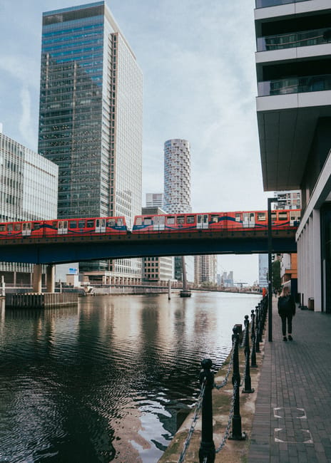 A view of a modern urban riverside scene with tall glass office buildings reflecting the sky and surrounding structures. A red train is crossing a bridge over the river, which has calm, reflective water. Along the right side, a paved walkway is bordered by black metal railings, with a person walking away from the camera. The scene is well-lit with natural daylight. Although the image primarily depicts city architecture and transportation, Canary Wharf Carpet Cleaning offers surface cleaning and deep cleaning services for office interiors and upholstery, ensuring hygiene and maintained appearance in commercial environments near Canary Wharf station. Visit canarywharfcarpetcleaning.org.uk for more information.