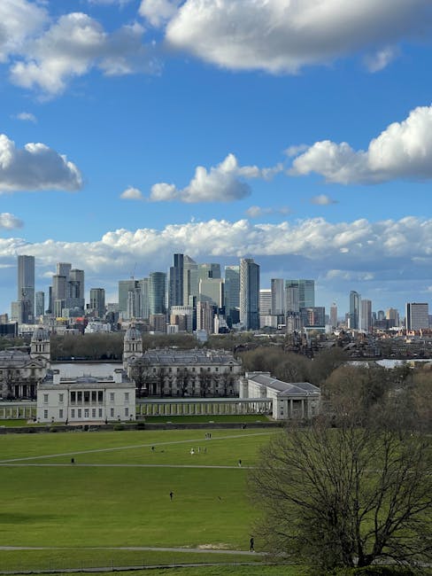 A wide view of Jubilee Park near Canary Wharf station showing historic neoclassical buildings with domed rooftops and ornate details, surrounded by green trees and open grassy areas. In the background, a dense cluster of modern skyscrapers with glass facades and rectangular shapes rise under a cloudy sky, creating a contrast between historic and contemporary architecture. The scene is outdoors during daylight, with some people walking or sitting on benches across the park, and a few vehicles visible near the buildings. The environment appears clean and well-maintained, typical of urban surface cleaning and civic space upkeep, as emphasized by Canary Wharf Carpet Cleaning in relation to detailed surface cleaning and hygiene.