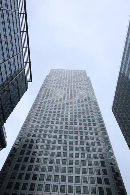 A view looking up at the exterior of a tall, modern skyscraper in Canary Wharf, featuring a grid of uniform windows along its glass and steel façade. Adjacent buildings with reflective glass surfaces frame the central tower, all under a cloudy, overcast sky. The scene highlights the sleek, clean lines of urban architectural surfaces, emphasizing the reflective glass and metal materials, with no visible dirt or debris. This image is relevant for content on surface cleaning and maintenance for commercial properties, as provided by Canary Wharf Carpet Cleaning, demonstrating the importance of hygiene and upkeep of high-rise surfaces.