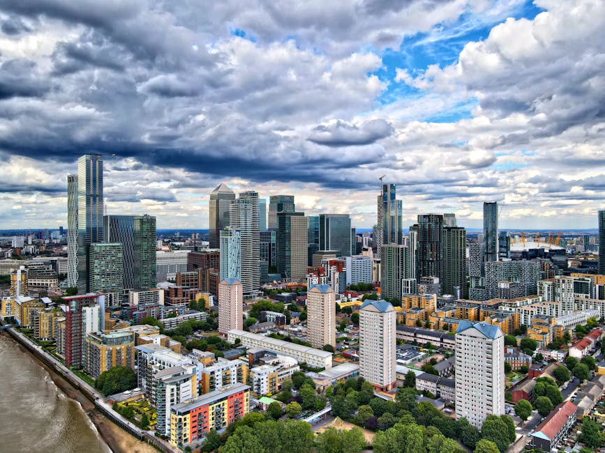 A high-angle view of the Canary Wharf commercial district in London, featuring a cluster of modern glass skyscrapers with reflective surfaces, surrounding mid-rise office buildings painted in pastel and neutral tones. The scene shows a mix of architectural styles, with some buildings having angular facades and others with curved or rectangular shapes. Green spaces with trees and small parks are interspersed among the structures. The river Thames runs along the bottom edge of the image, with a paved walkway bordering the riverbank. The sky is filled with dense, partly cloudy clouds allowing patches of blue sky to peek through, creating a dynamic lighting contrast over the cityscape. The overall scene appears clean, well-maintained, and vibrant, illustrating the urban environment in the vicinity of Canary Wharf as seen from an elevated vantage point.
