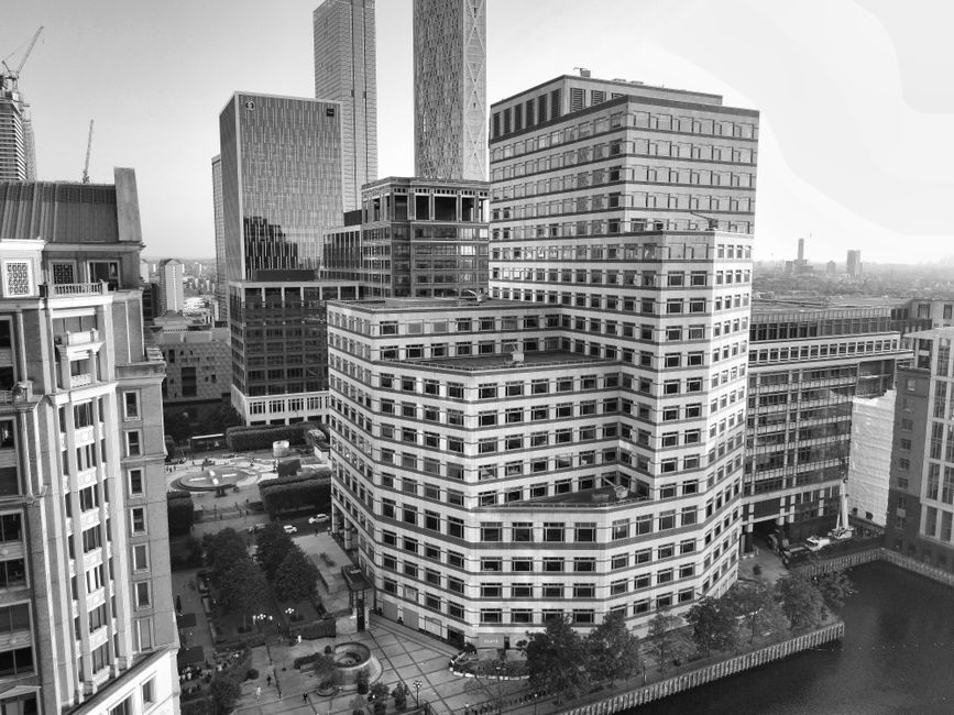 Black and white aerial photograph of a cluster of modern high-rise office buildings in Canary Wharf, featuring distinctive architectural designs with numerous windows and flat rooftops. The buildings are arranged around a landscaped courtyard with pathways, small trees, and a few parked cars visible at ground level. The scene is well-lit with natural daylight broadening the view of the surrounding cityscape, highlighting reflections on glass facades. The image exemplifies urban surface and facade details associated with commercial architecture, aligning with the focus of South Quay emergency carpet cleaning services by Canary Wharf Carpet Cleaning for maintaining hygiene and cleanliness in large-scale office environments.