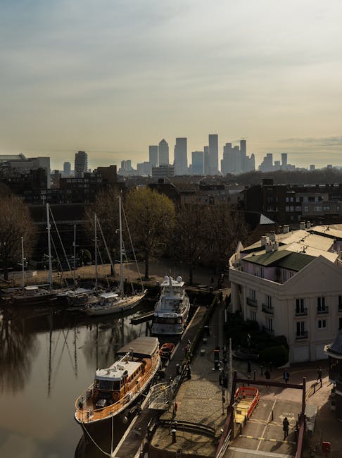 A high-angle view of the Canary Wharf commercial district in London, featuring a cluster of modern glass skyscrapers with reflective surfaces, surrounding mid-rise office buildings painted in pastel and neutral tones. The scene shows a mix of architectural styles, with some buildings having angular facades and others with curved or rectangular shapes. Green spaces with trees and small parks are interspersed among the structures. The river Thames runs along the bottom edge of the image, with a paved walkway bordering the riverbank. The sky is filled with dense, partly cloudy clouds allowing patches of blue sky to peek through, creating a dynamic lighting contrast over the cityscape. The overall scene appears clean, well-maintained, and vibrant, illustrating the urban environment in the vicinity of Canary Wharf as seen from an elevated vantage point.